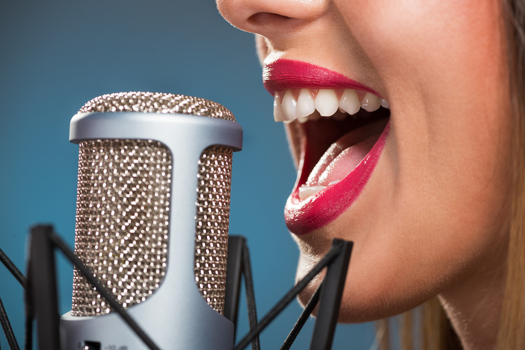 Female with pink lipstick singing into a microphone against a blue backdrop. 