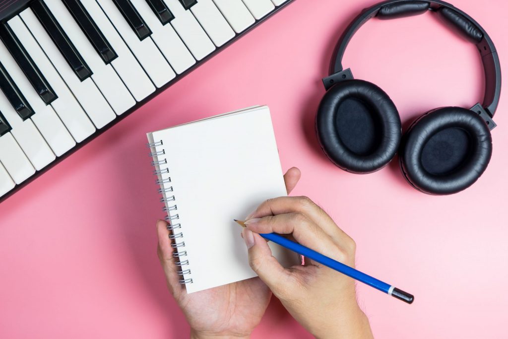 Pink desktop with a keyboard piano on the left, headphones on the right, and a woman's hands writing into a journal with a blue pencil in her right hand.