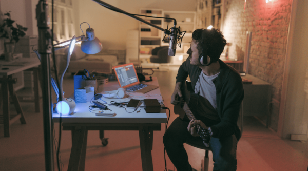 Guy playing guitar sitting in front of his desk with a microphone above and laptop and notes on his desk