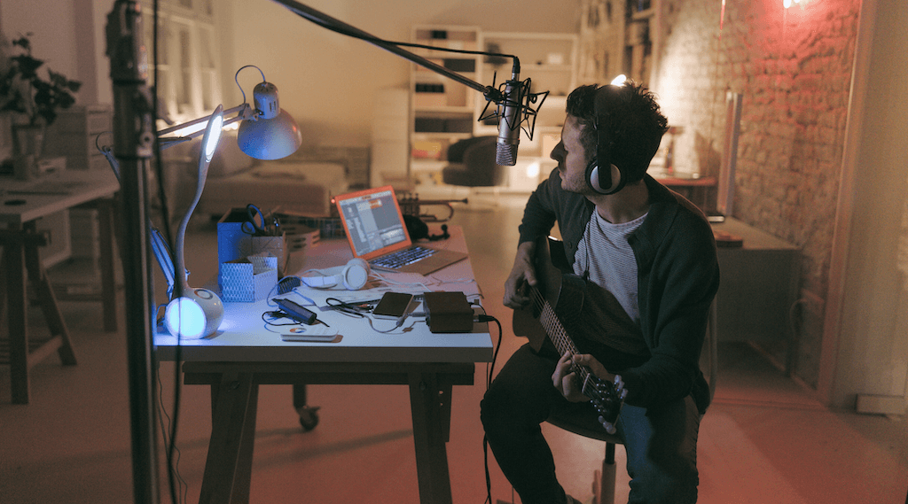  Guy playing guitar sitting in front of his desk with a microphone above and laptop and notes on his desk