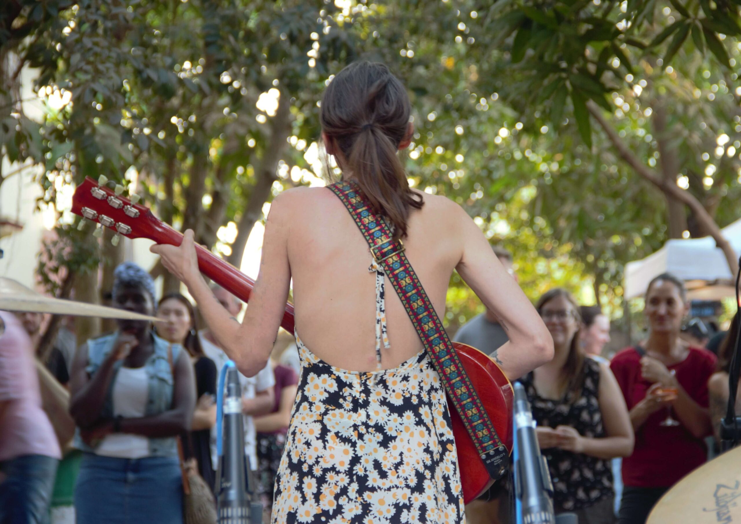 Girl playing a reddish brown guitar looking out toward audience outside under the trees