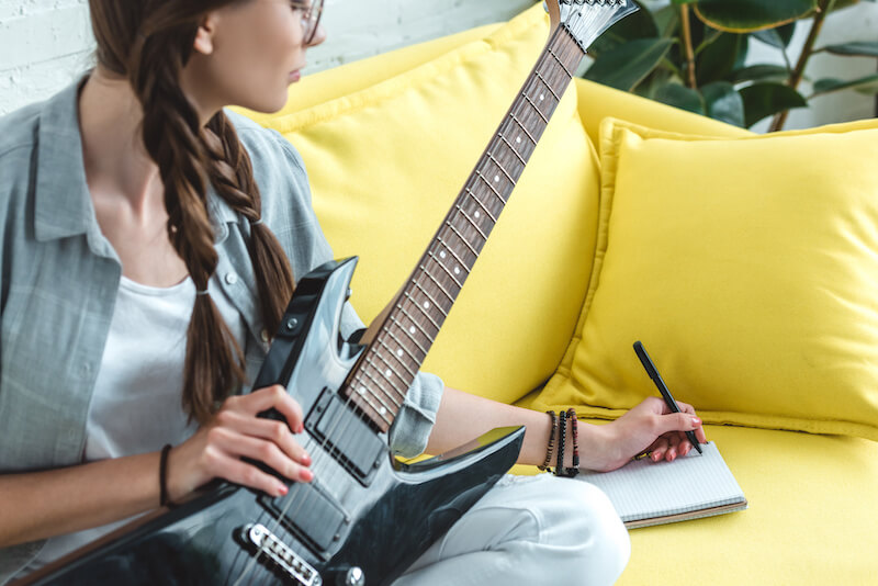 Woman holding an electric guitar sitting on a yellow couch and writing in a journal