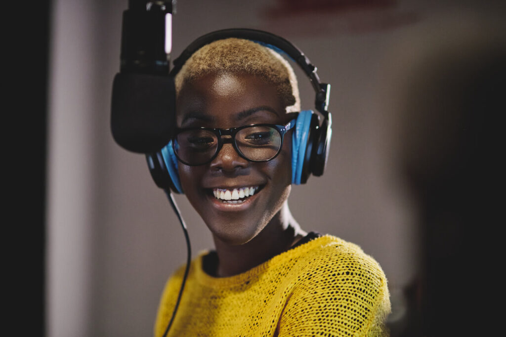 Woman wearing headphones and a yellow sweater while smiling in front of a microphone.