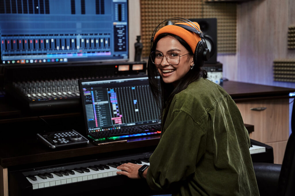 Female musician with black hair wearing a green jacket, orange beanie, and headphones while mixing music in the recording studio.