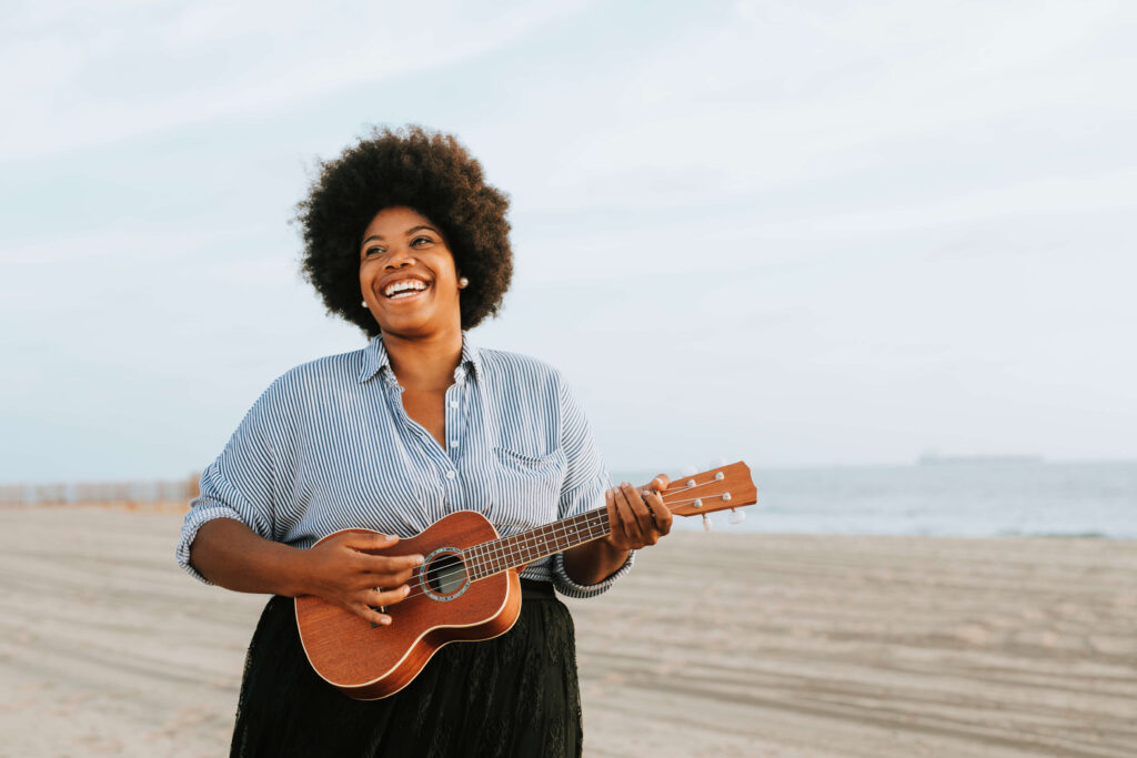 Black female musician smiling, wearing a black dress with a blue and white striped shirt while playing a ukulele at the beach.