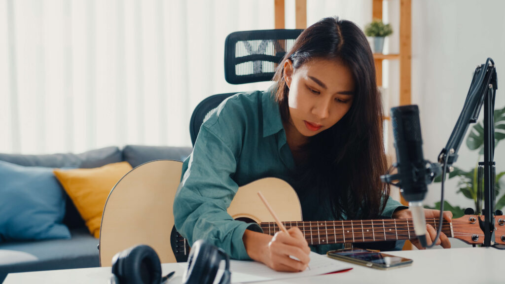 Female musician with black hair sitting in her home studio sitting at a desk with microphone while holding a guitar and writing in a notebook. 