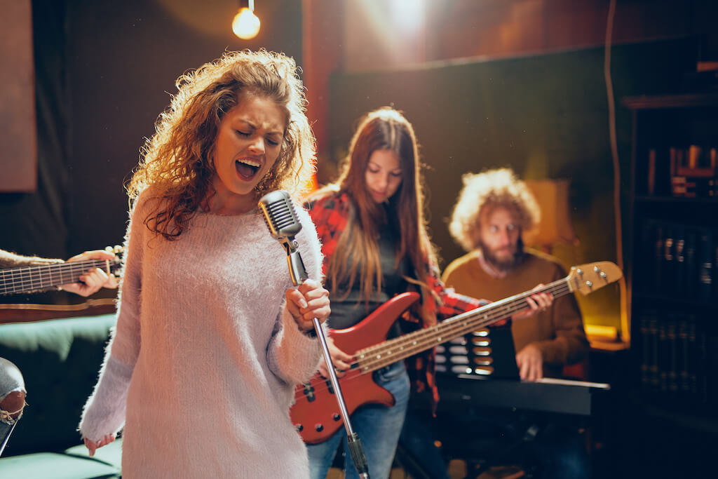 Female singer with curly blonde hair singing into a microphone while her band behind her plays with her