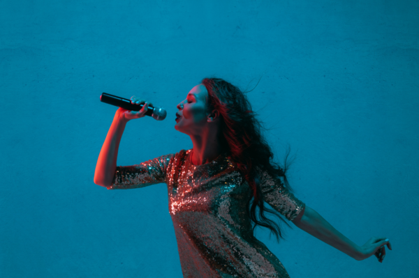 female musician with long brown hair and silver sequined dress singing into a microphone with plain blue background