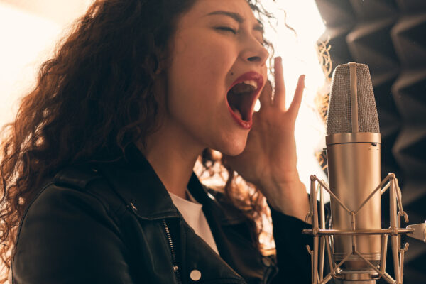 Portrait of beautiful curly woman in leather jacket and hat recording vocals in music studio on professional sound equipment