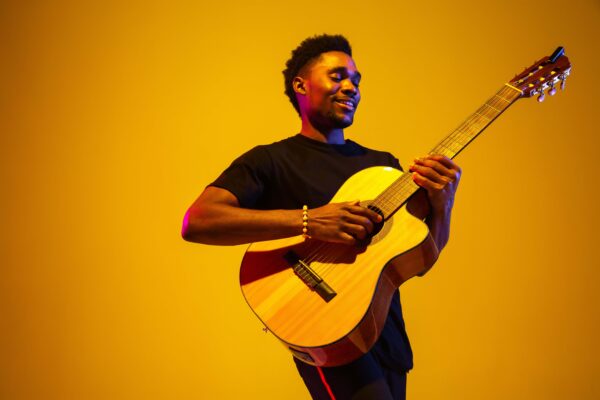 A smiling man playing an acoustic guitar against a bright yellow background, wearing a black shirt and bracelet.