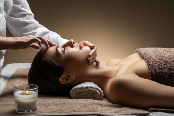 A woman lying on a massage table with her eyes closed, receiving a relaxing facial and head massage from a therapist. A lit candle sits nearby, creating a calm and soothing atmosphere.